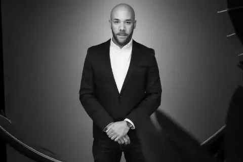 Portrait noir et blanc d’un homme en costume sombre et chemise blanche, mains jointes.