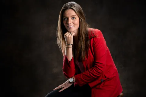 Femme en blazer rouge, main sous le menton, sur fond brun texturé.
