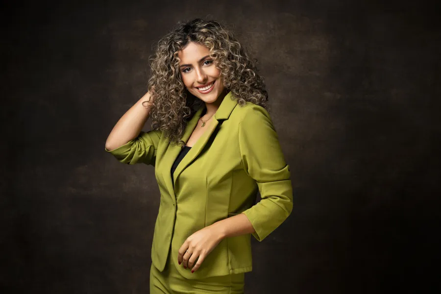 Femme en costume vert anis, main dans les cheveux, sur fond brun.