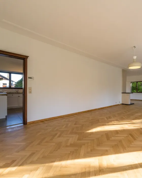 Grande pièce vide baignée de soleil, parquet en chevrons et portes ouvertes en enfilade.