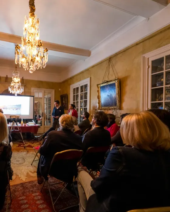 Conférence dans un salon de manoir sous un lustre, public attentif face à l’écran de présentation.