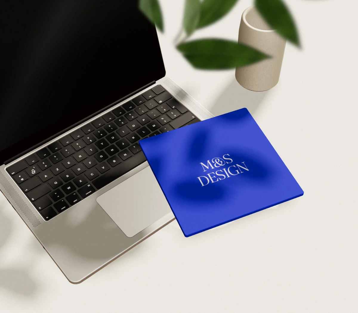 Un ordinateur portable ouvert sur un bureau clair, un carnet bleu M&S Design posé dessus, l’ombre d’une plante.