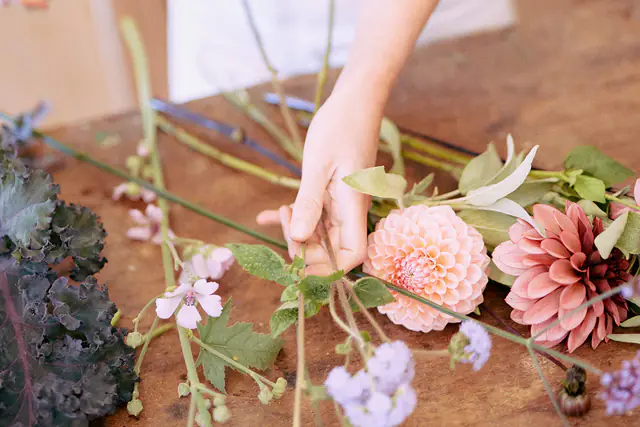 Handen die een boeket met dahlia’s en seizoensbloemen schikken op de houten tafel van het atelier